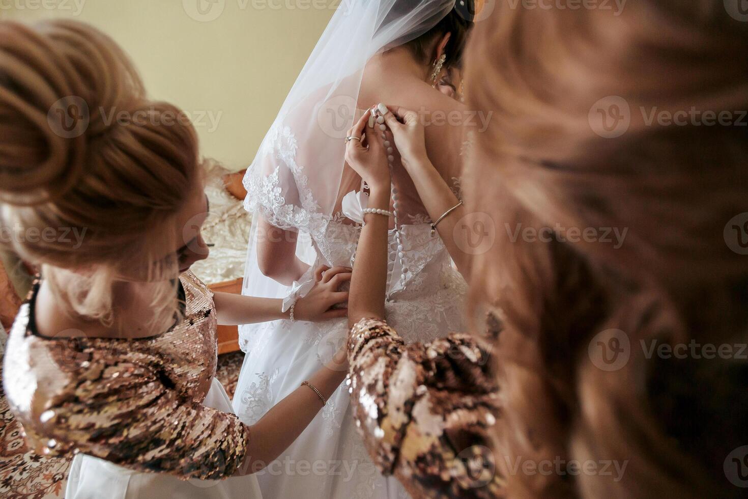 Three women are getting ready for a wedding. One of them is adjusting the bride's dress. The other two women are standing behind her, watching and helping photo