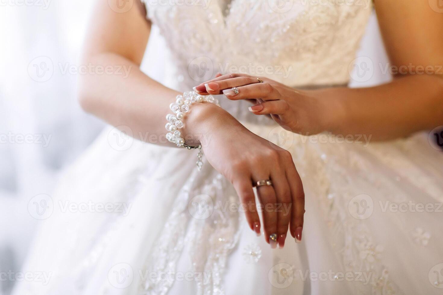 A woman in a white dress is wearing a pearl bracelet. She is standing in front of a window, and her hands are clasped together. Concept of elegance and sophistication photo