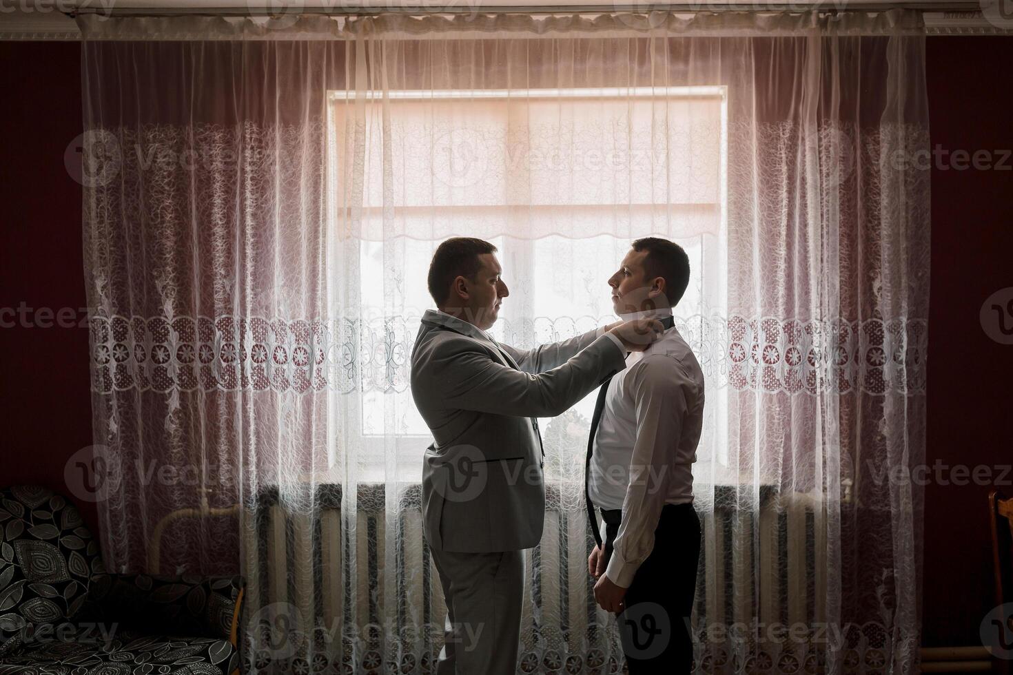 Two men are getting dressed in front of a window. One man is helping the other tie his tie photo