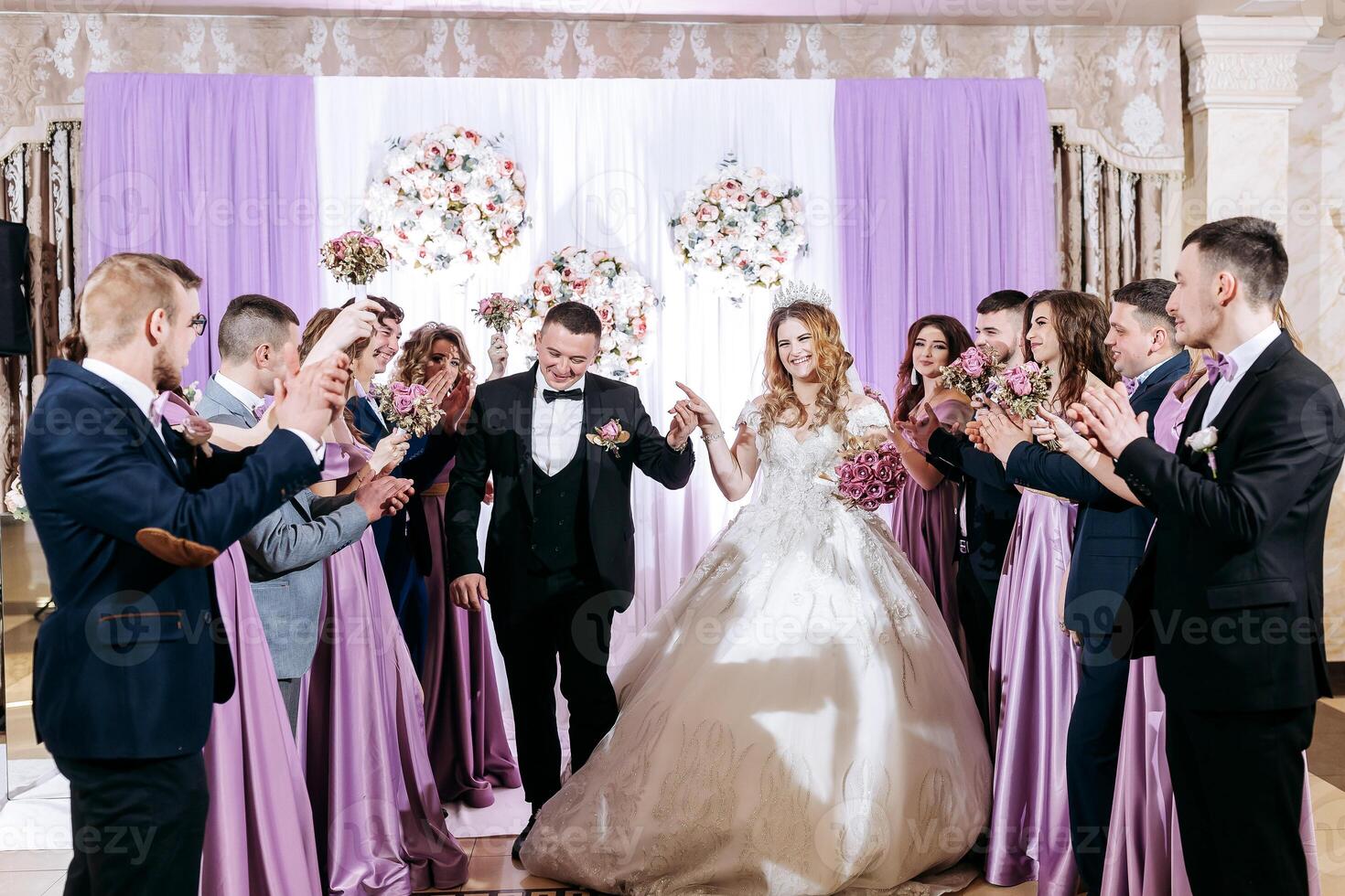 A bride and groom are walking down a purple aisle with a group of people clapping. The bride is wearing a long white dress and the groom is wearing a black suit photo