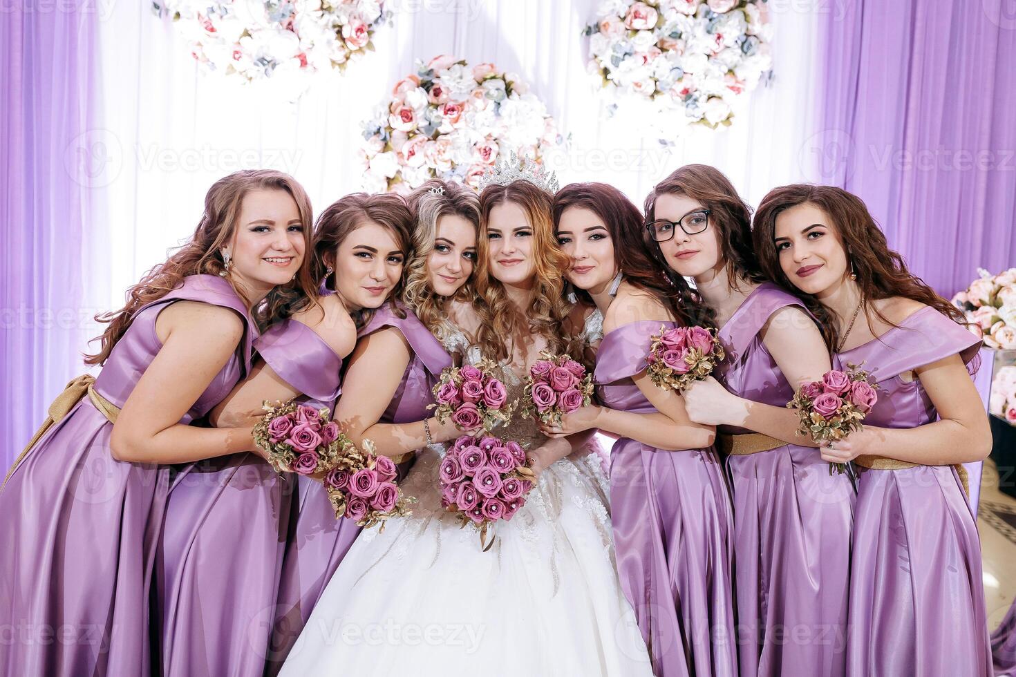 A group of women are posing for a picture, with one of them holding a bouquet of flowers. The bride is wearing a white dress and is surrounded by her bridesmaids in purple dresses photo
