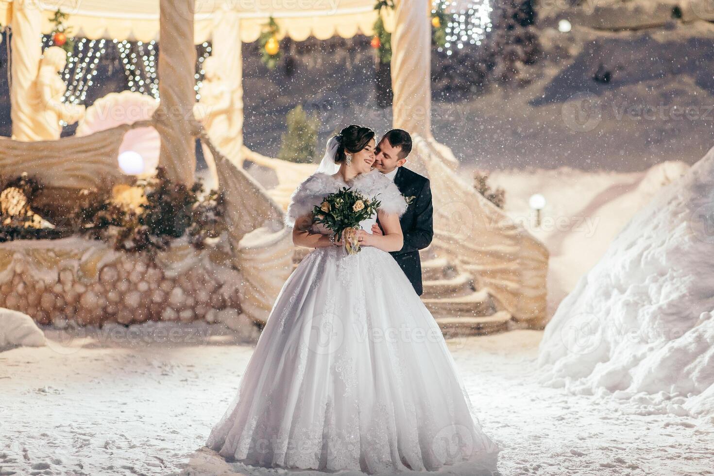 A bride and groom are standing in the snow, with the bride wearing a white dress and the groom wearing a black suit. They are holding hands and smiling, creating a warm and happy atmosphere photo