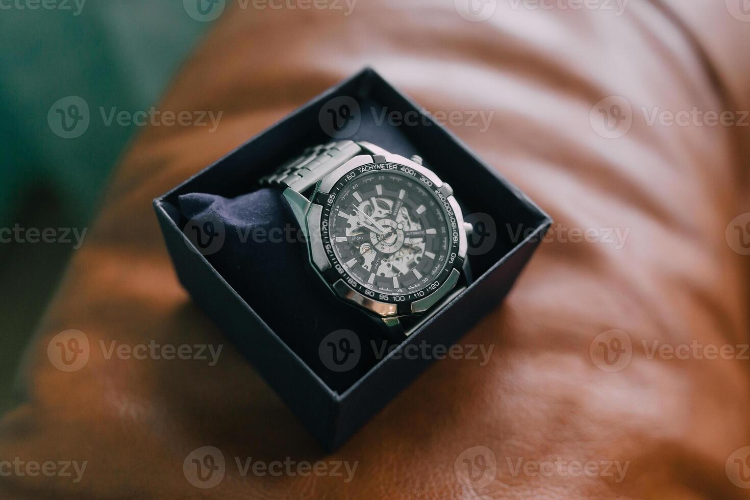 A watch is sitting in a box on a brown surface. The watch is silver and has a black face photo