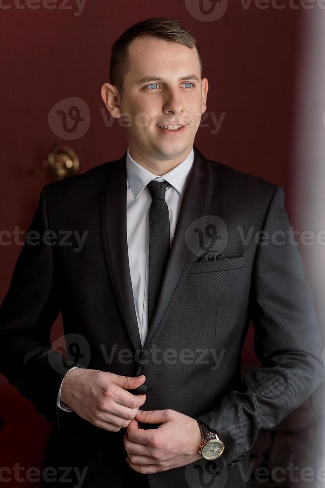 A man in a suit and tie is standing in front of a red wall. He is wearing a watch and has a smile on his face photo