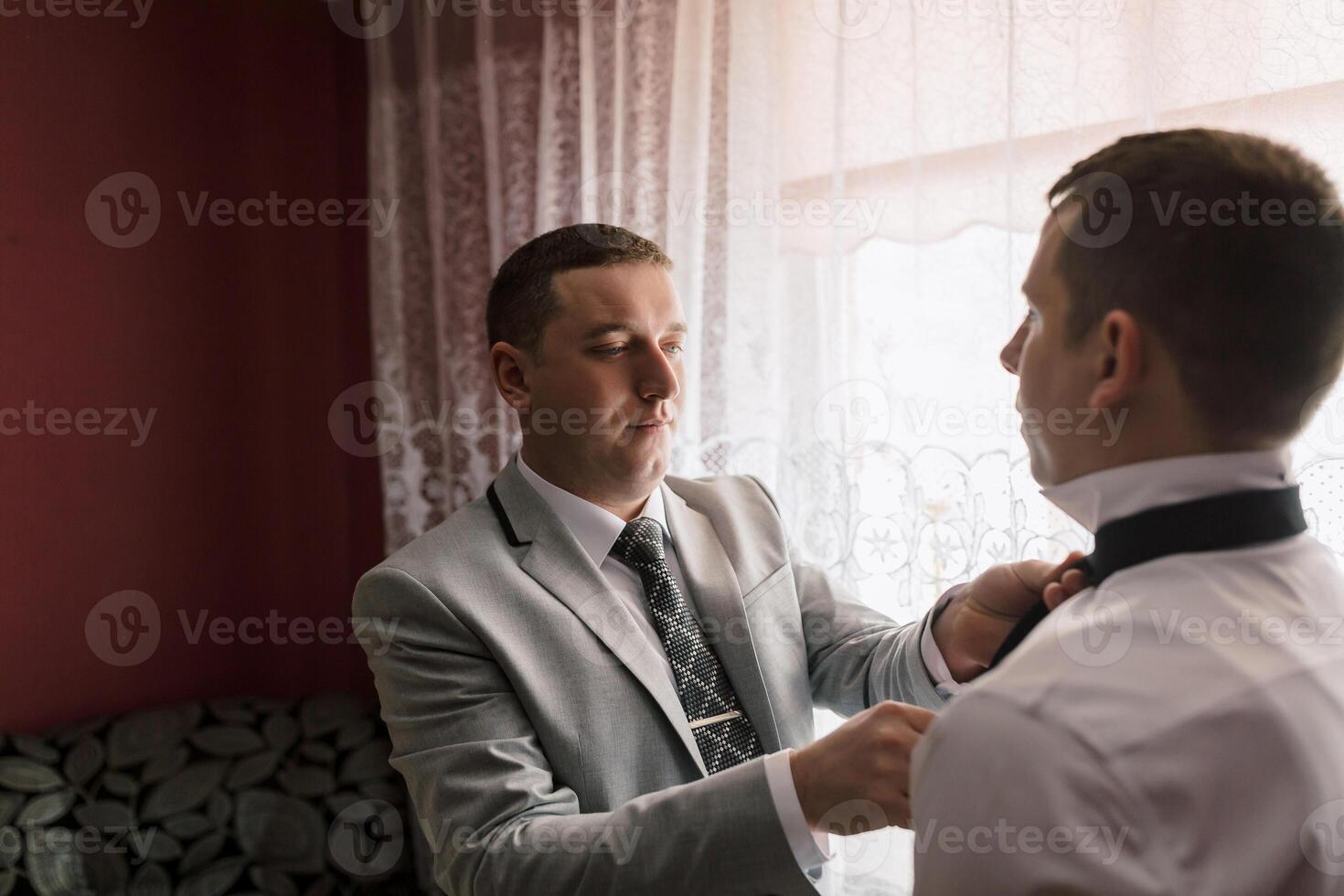 Two men are getting dressed up, one of them is adjusting the tie of the other. The man adjusting the tie is wearing a gray suit and tie, while the other man is wearing a white shirt photo