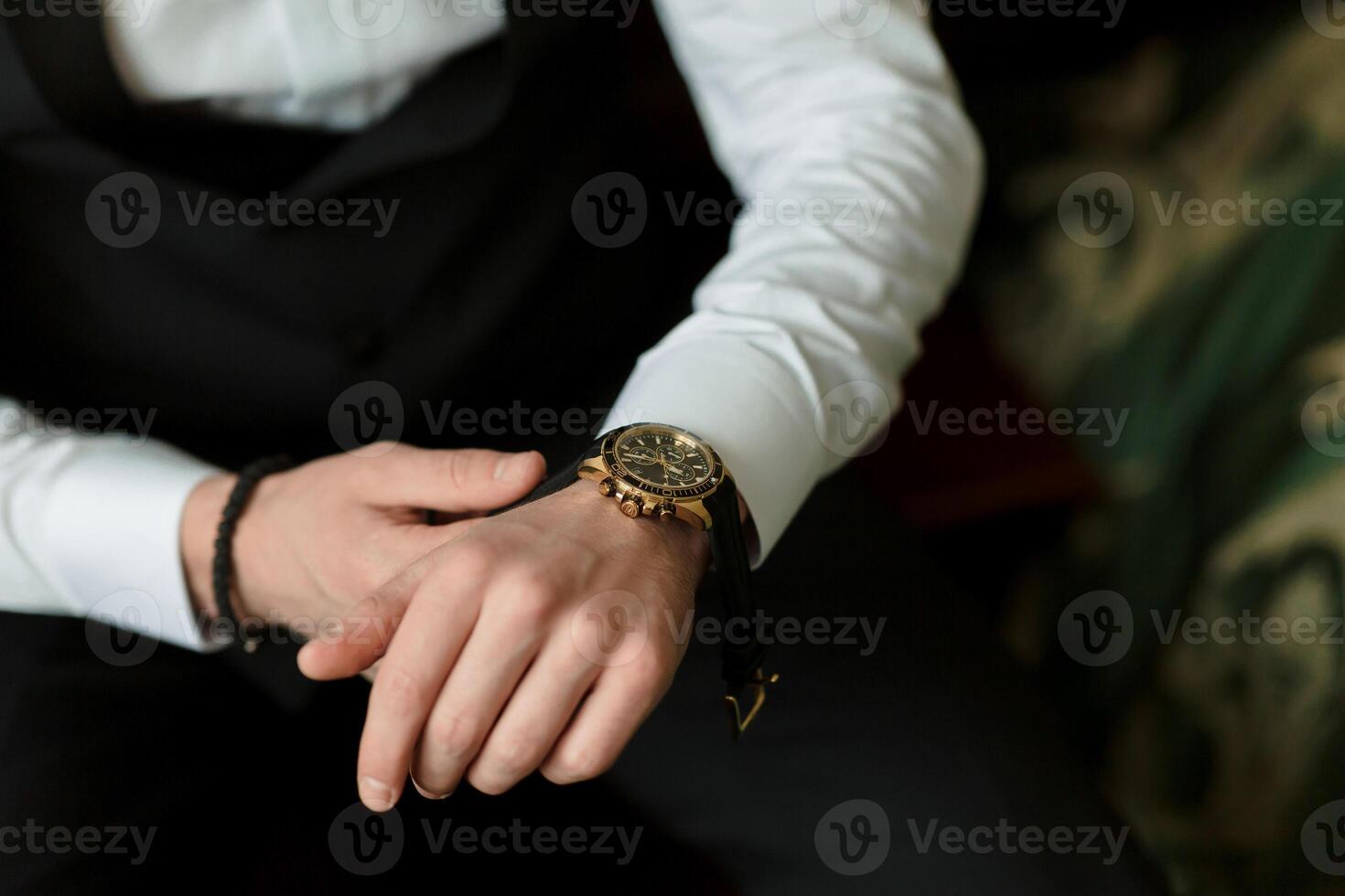 A man is wearing a black suit and a gold watch. He is sitting on a chair and looking at his watch photo