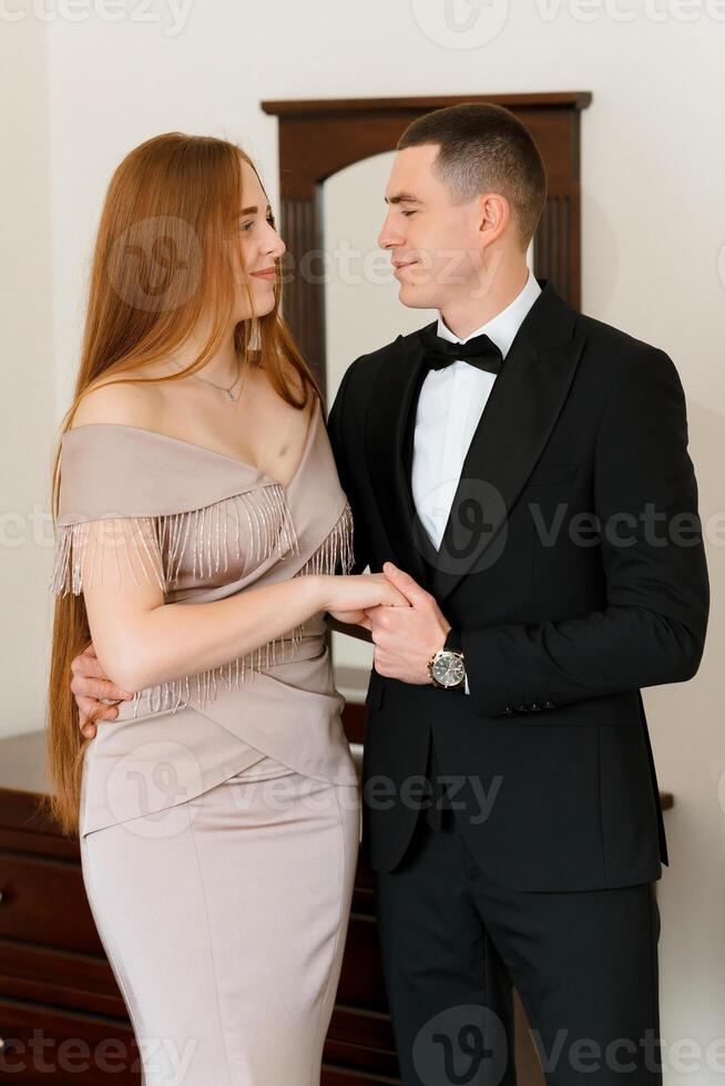 A man and a woman are standing in front of a mirror, both wearing formal attire. The man is wearing a black suit and a white tie, while the woman is wearing a dress with a long train photo