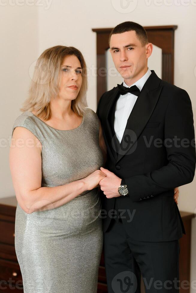 A woman and a man are posing for a picture. The woman is wearing a silver dress and the man is wearing a black suit. Scene is formal and elegant photo