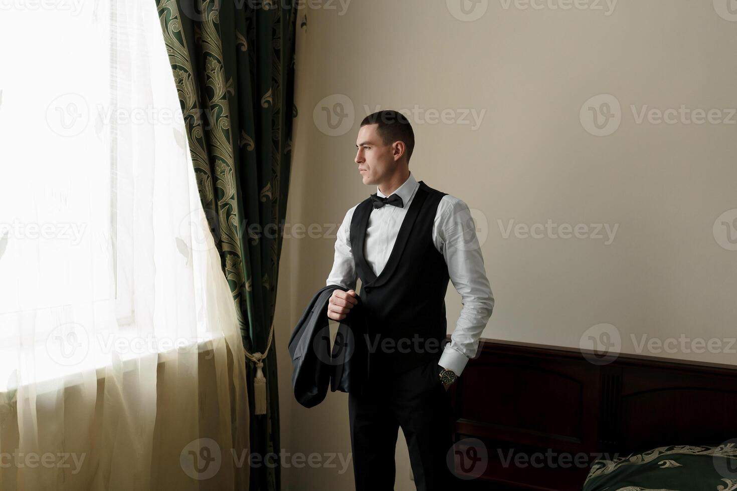 A man in a black vest and a black bow tie is standing in front of a window. He is holding a black briefcase in his hand. Concept of formality and professionalism photo