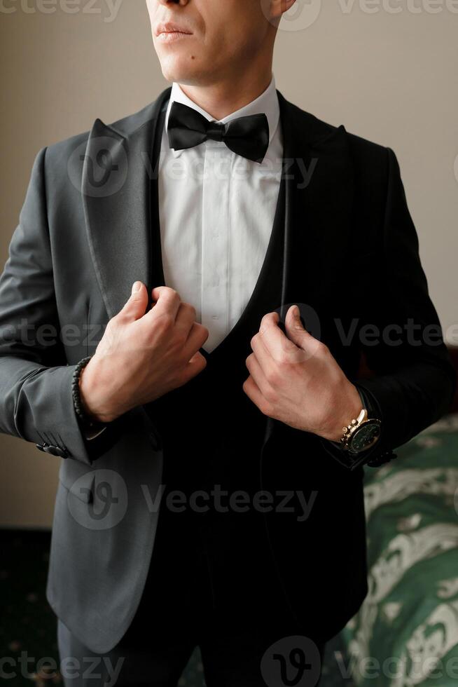A man in a black suit and white shirt is getting ready to wear a black bow tie. He is adjusting his suit jacket and tie, and his hands are visible. Concept of formality and elegance photo