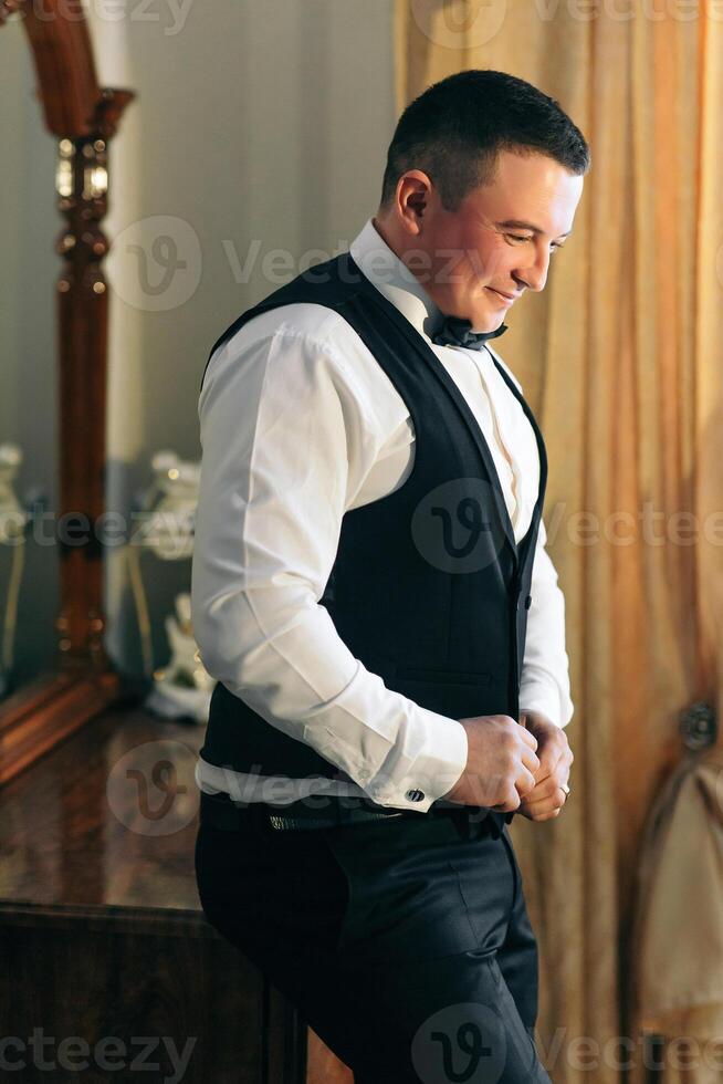 A man in a black vest and white shirt is standing in front of a mirror. He is adjusting his tie and he is in a good mood photo