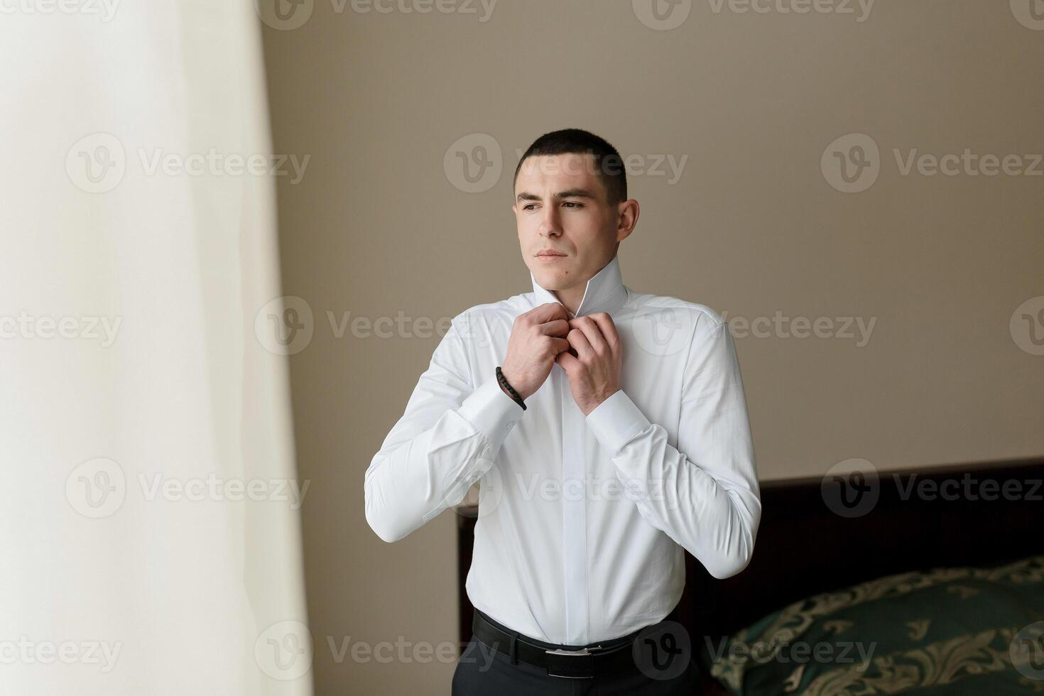 A man in a white shirt is getting dressed and adjusting his collar. Concept of formality and attention to detail, as the man takes care to ensure his appearance is neat and polished. The setting photo