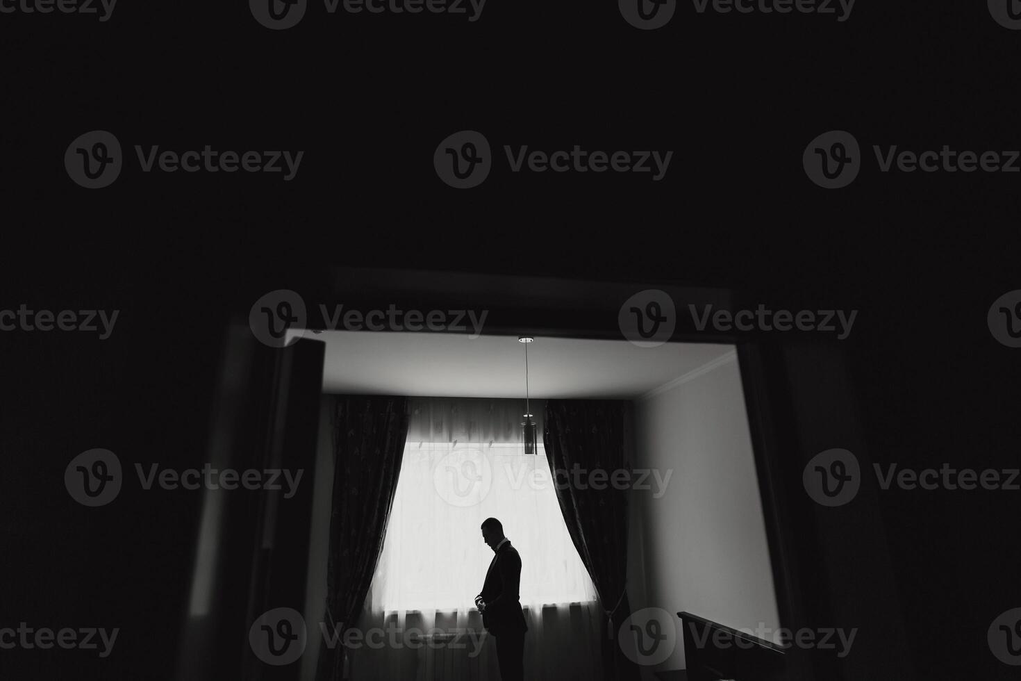 A man is standing in a room with a window. He is wearing a suit and he is pregnant. The room is dark and the man is the only person in it photo