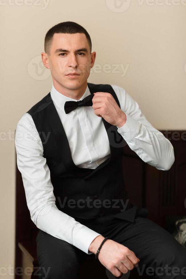 A man in a black vest and white shirt is sitting on a bed. He is adjusting his tie and looking at the camera. Concept of formality and elegance, as the man is dressed in a suit and tie photo