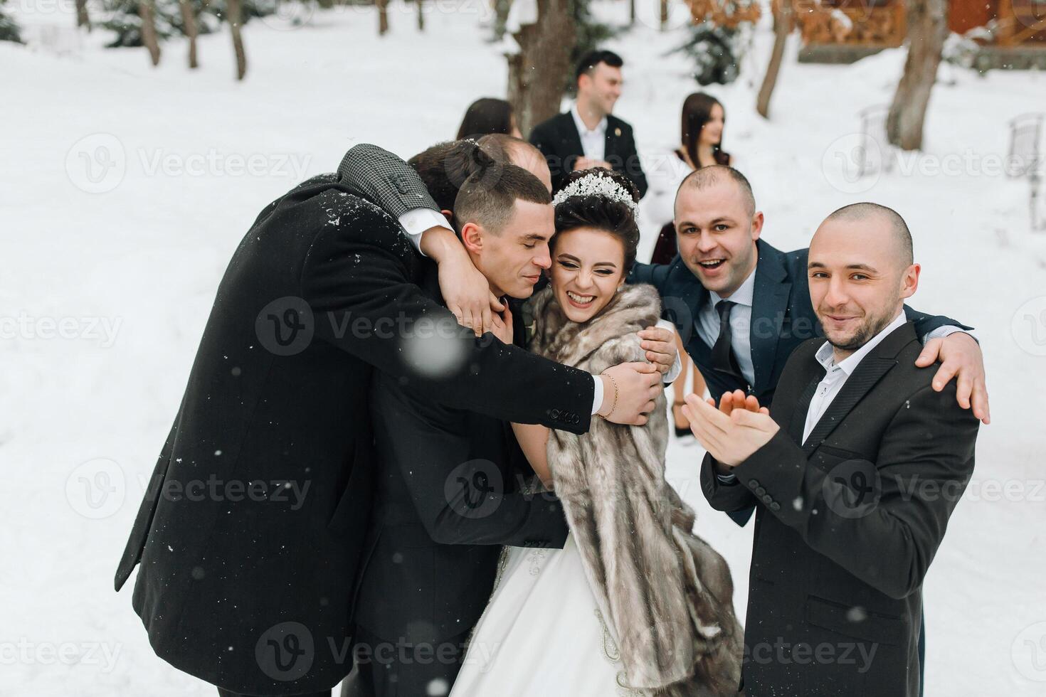 A bride and groom are surrounded by their friends and family, all of whom are smiling and hugging. The bride is wearing a fur stole and the groom is wearing a tie. Scene is joyful and celebratory photo