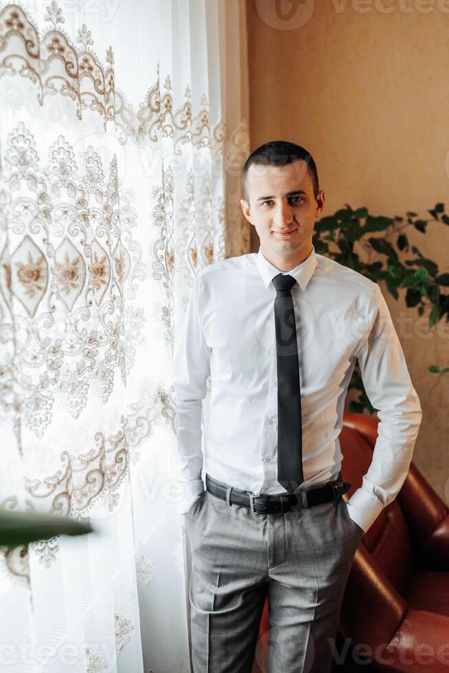 A man in a white shirt and gray pants stands in front of a window with a plant behind him. He is wearing a black tie and has his hands in his pockets. Concept of professionalism and formality photo