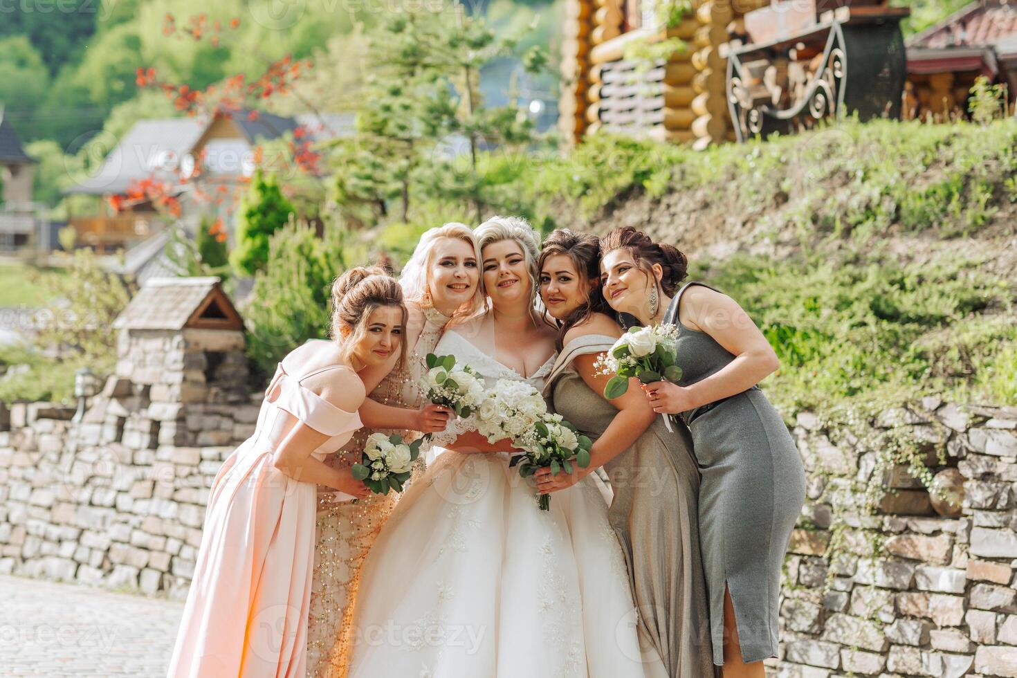A group of women are posing for a photo in front of a house. The bride is wearing a white dress and holding a bouquet of flowers. The other women are dressed in elegant gowns
