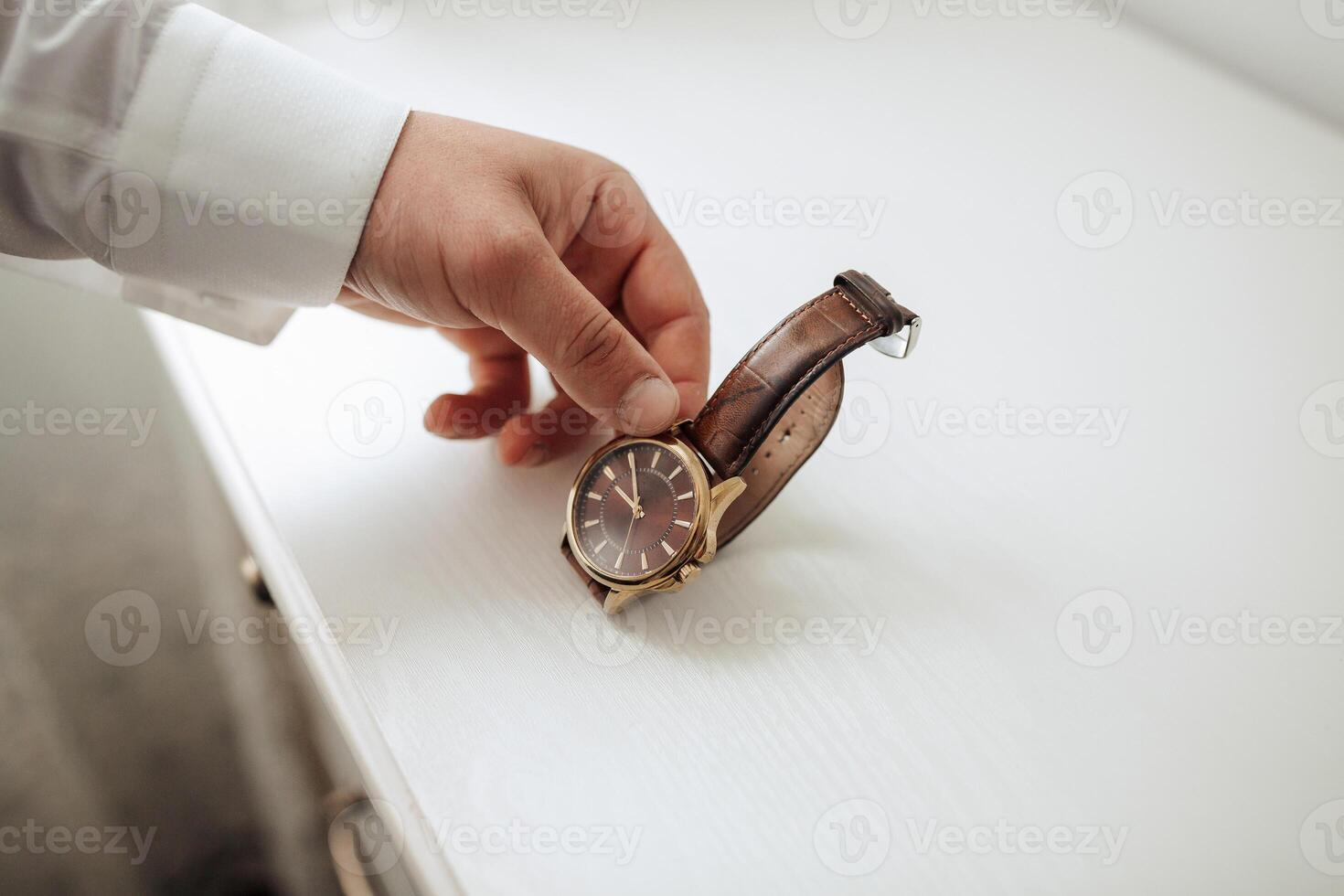 A man is holding a watch with a brown band. The man is wearing a white shirt photo