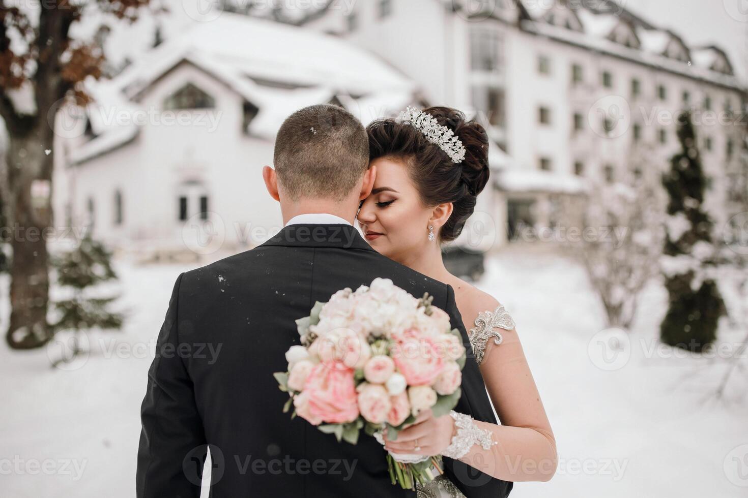 A bride and groom are embracing in the snow, with the bride holding a bouquet. The scene is set in a snowy landscape, with a large building in the background. Scene is romantic and intimate photo