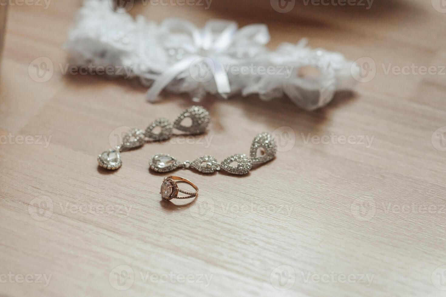 A wedding ring and a pair of earrings are displayed on a wooden table. The earrings are made of diamonds and are placed on top of the ring photo