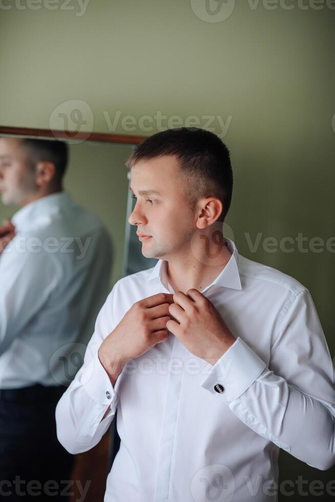 A man is getting dressed in front of a mirror. He is wearing a white shirt and a black suit photo
