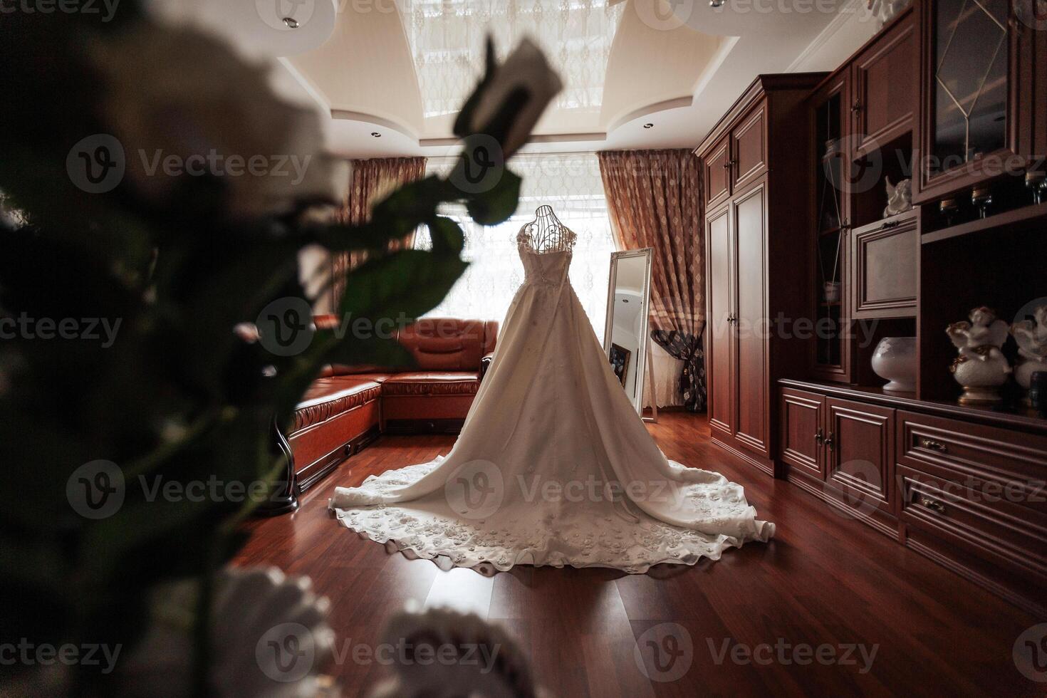 A bride's wedding dress is displayed in a room with a floral arrangement in the background. The dress is long and elegant, and the room is decorated with wooden furniture and a vase photo