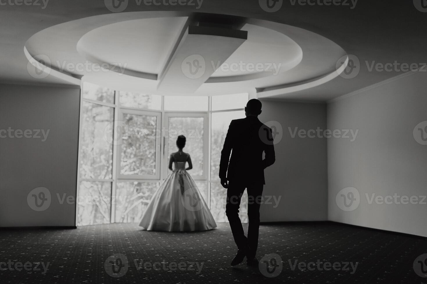 A man and a woman are standing in a room with a window. The man is wearing a suit and the woman is wearing a dress. Scene is romantic and intimate photo