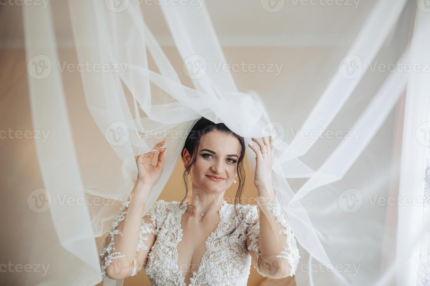 A bride is wearing a veil and standing in front of a curtain. She is smiling and looking at the camera photo