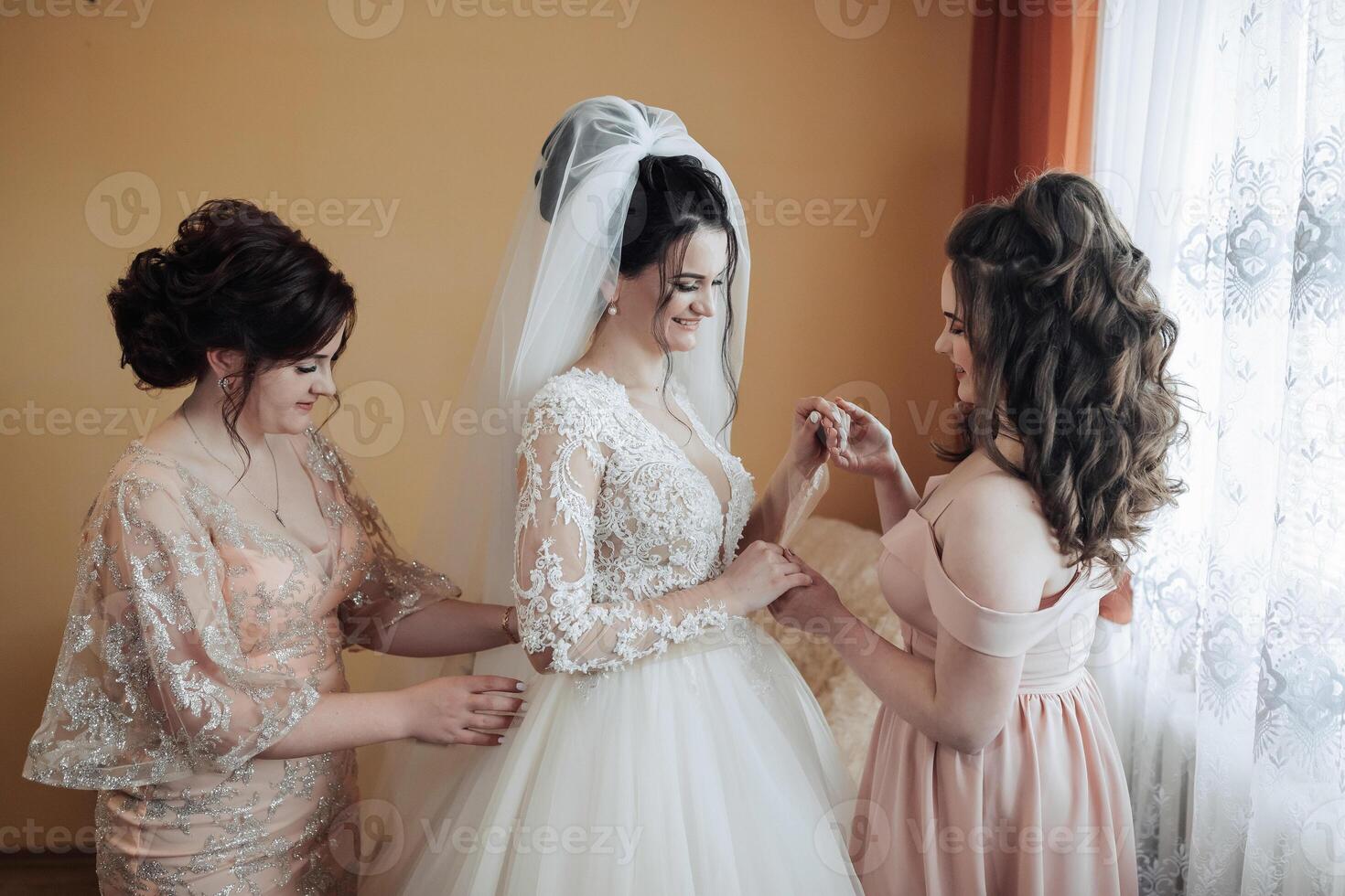 Three women are getting ready for a wedding. One of them is wearing a wedding dress and the other two are helping her with her dress photo