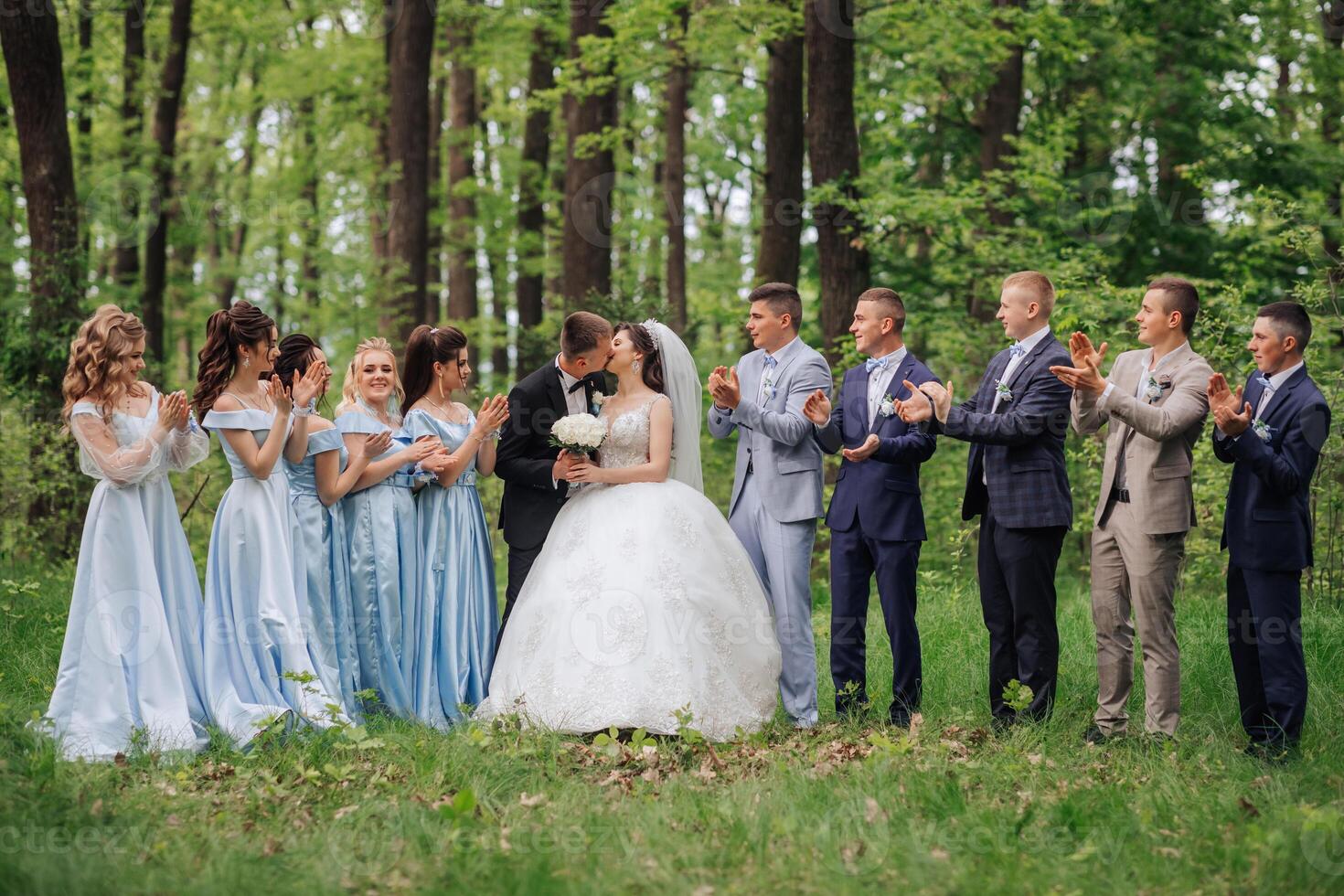 A group of people are standing in a field, with a bride and groom in the center. The bride is wearing a white dress and the groom is kissing her. The group is clapping and cheering for the newlyweds photo
