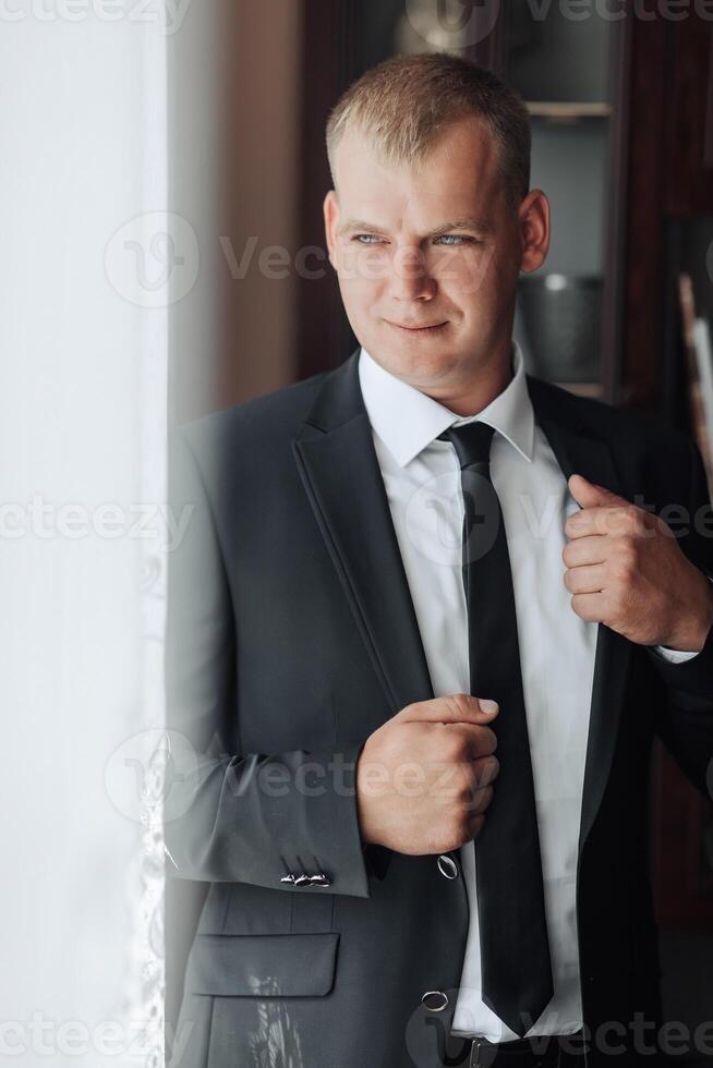 A man in a suit and tie is standing in front of a window, adjusting his tie. Concept of formality and professionalism, as the man is dressed in a suit and tie photo
