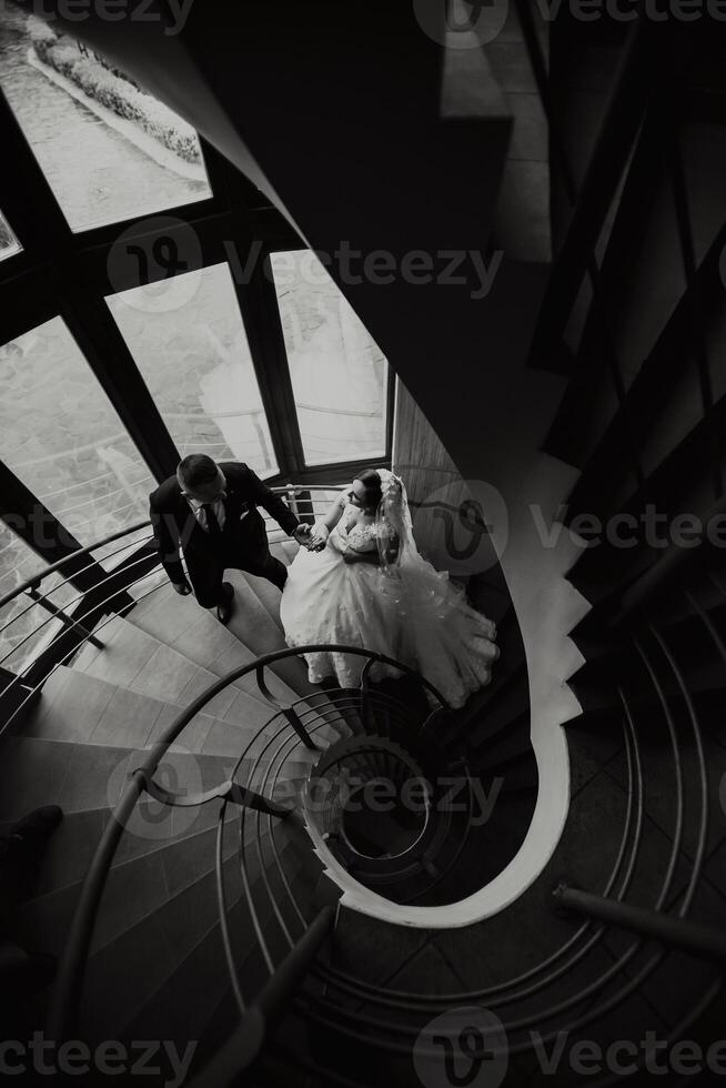 A bride and groom are walking up a spiral staircase. The bride is wearing a white dress and the groom is wearing a suit. Concept of romance and love, as the couple is dressed up for their wedding day photo