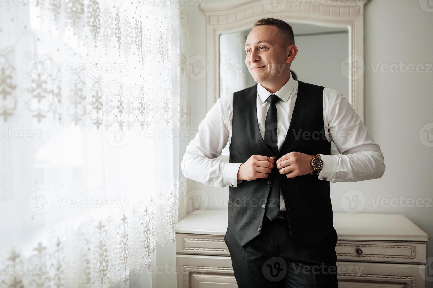 A man in a suit is standing in front of a window, adjusting his tie. He is wearing a black vest and a white shirt photo