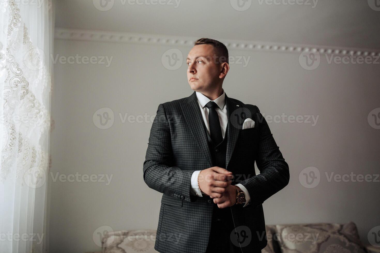 A man in a suit is standing in front of a window, adjusting his tie. Concept of formality and professionalism, as the man is dressed in a suit and tie. The window provides a view of the outside world photo