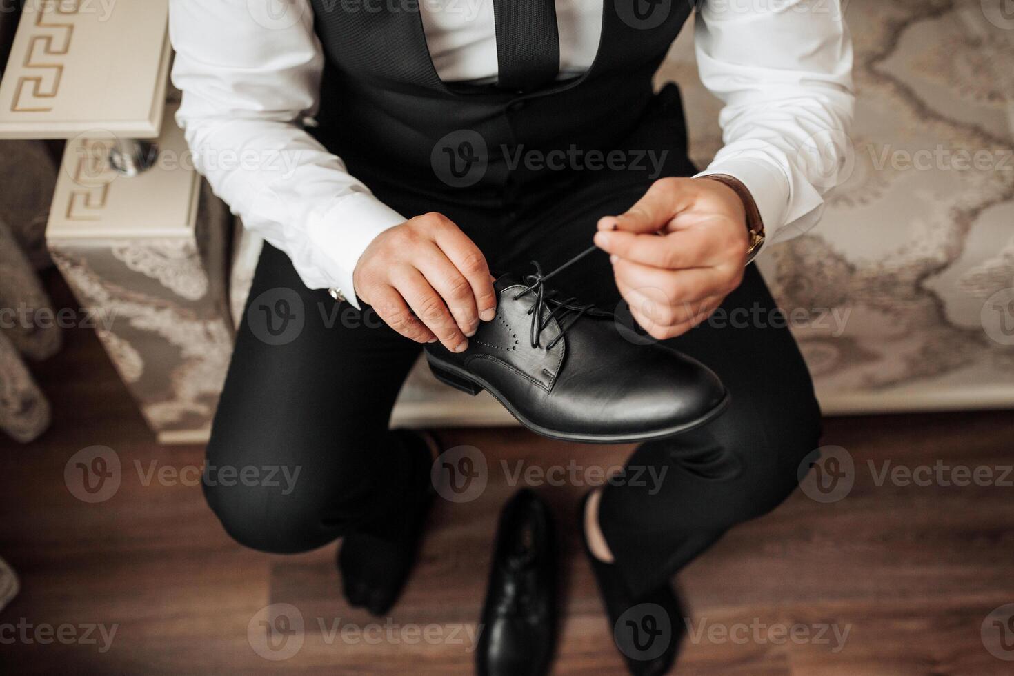 A man in a suit is putting on his shoes. He is sitting on a wooden floor. The shoes are black and shiny photo