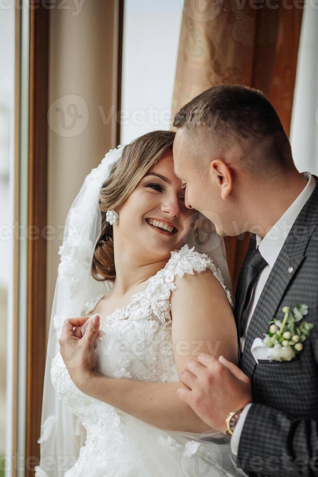 A bride and groom are hugging and smiling at the camera. The bride is wearing a white dress and the groom is wearing a suit. Scene is happy and joyful, as the couple is celebrating their wedding day photo