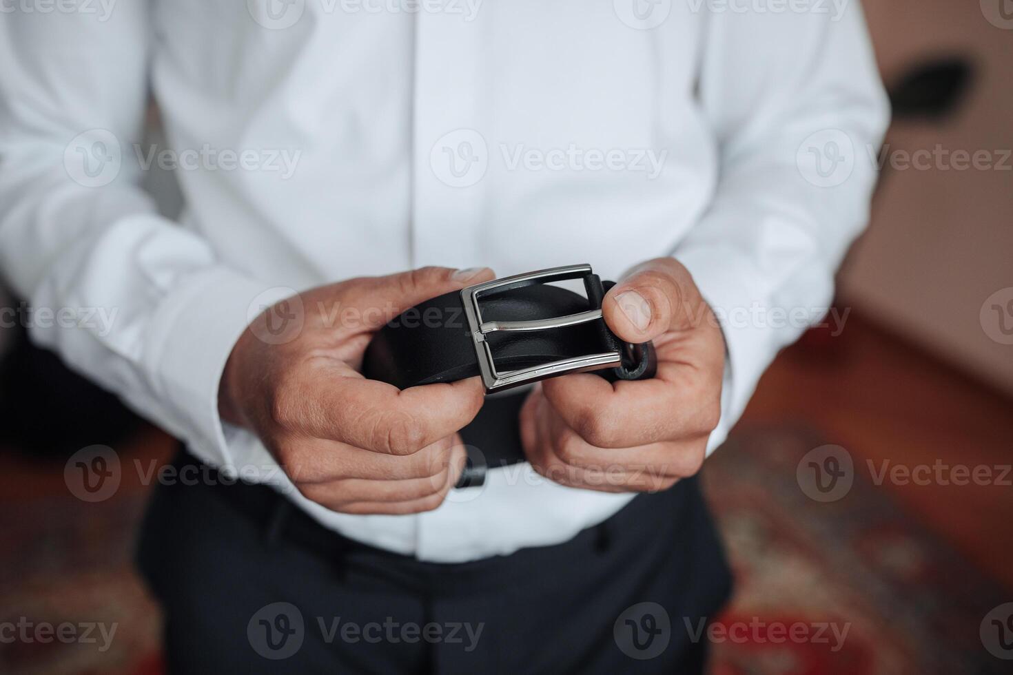 A man is holding a belt in his hand. The belt is black and has a silver buckle. The man is wearing a white shirt and black pants. Concept of formality and elegance photo