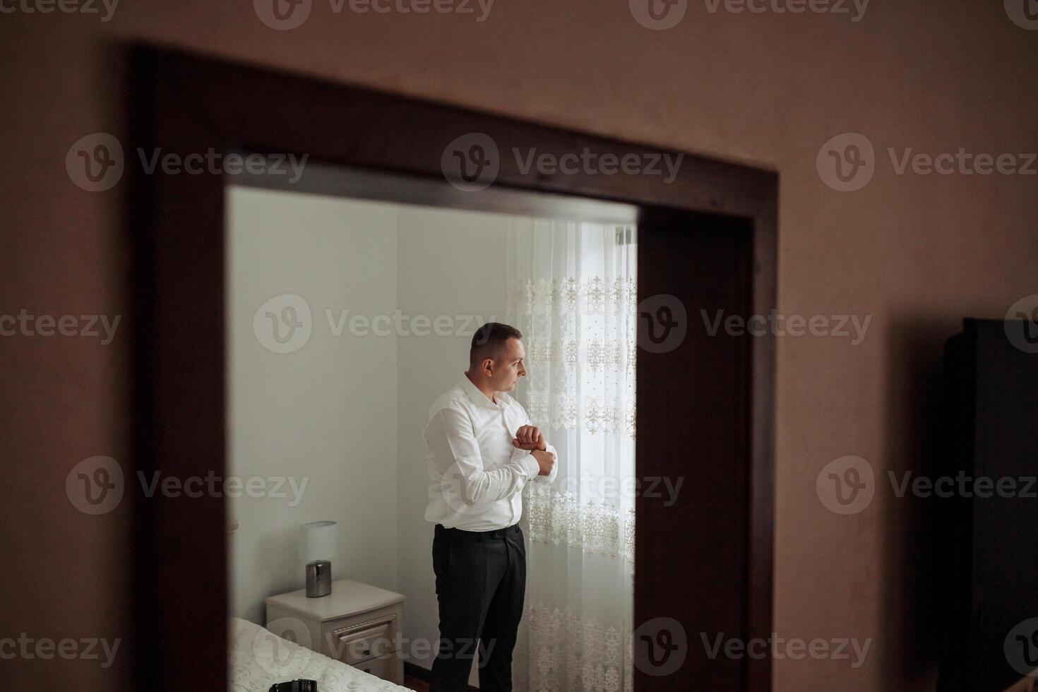 A man is standing in front of a mirror, getting ready for the day. He is wearing a white shirt and black pants photo