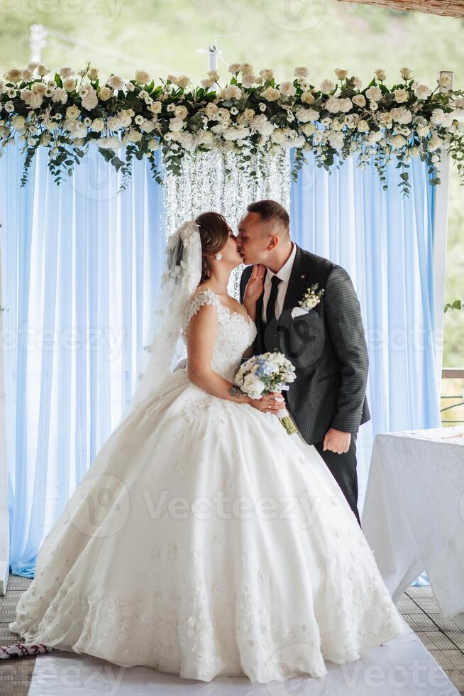 A bride and groom kiss in front of a blue curtain. The bride is wearing a white dress and the groom is wearing a suit photo