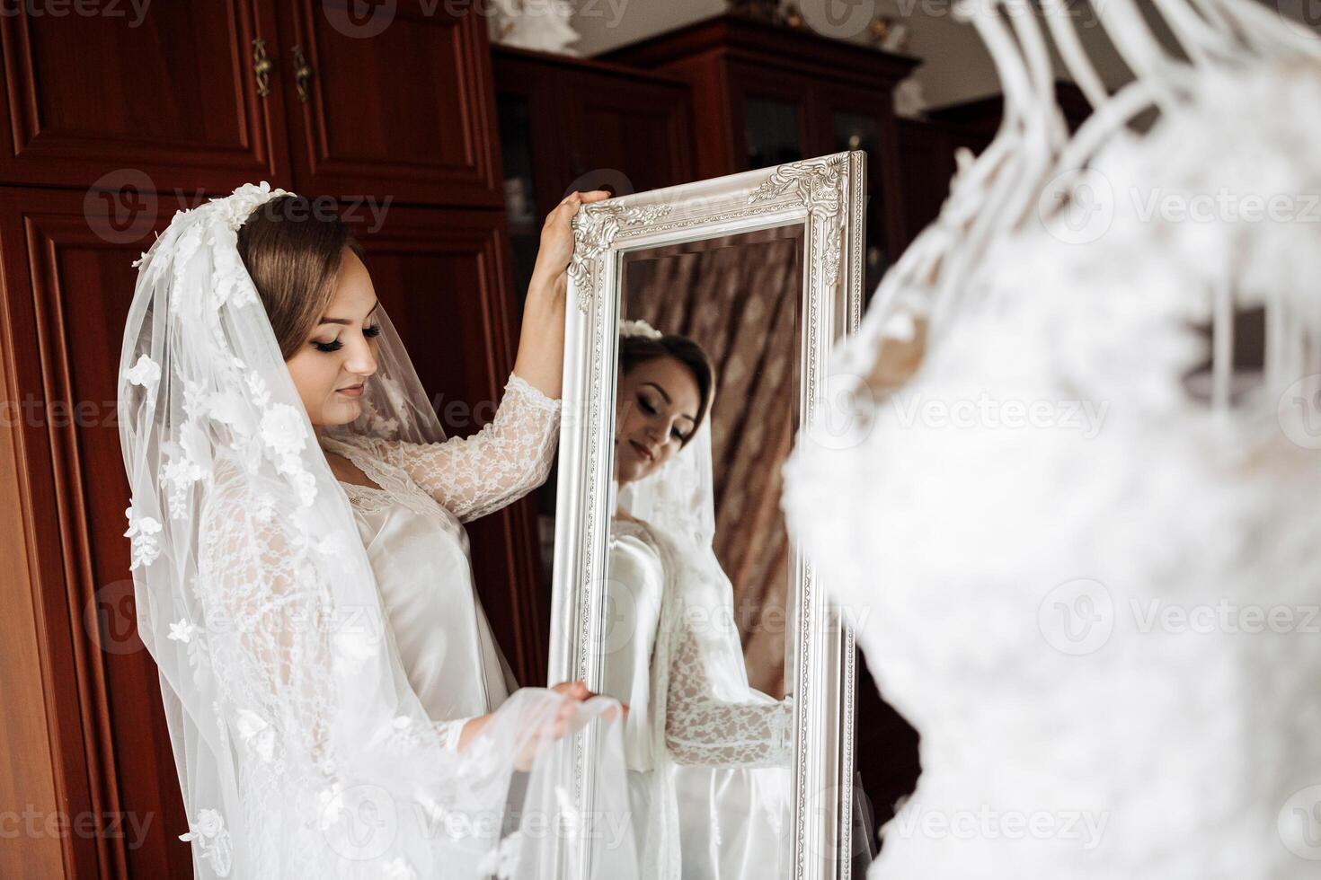 A woman is getting ready for her wedding and is looking at herself in a mirror. The mirror is next to a white dress photo