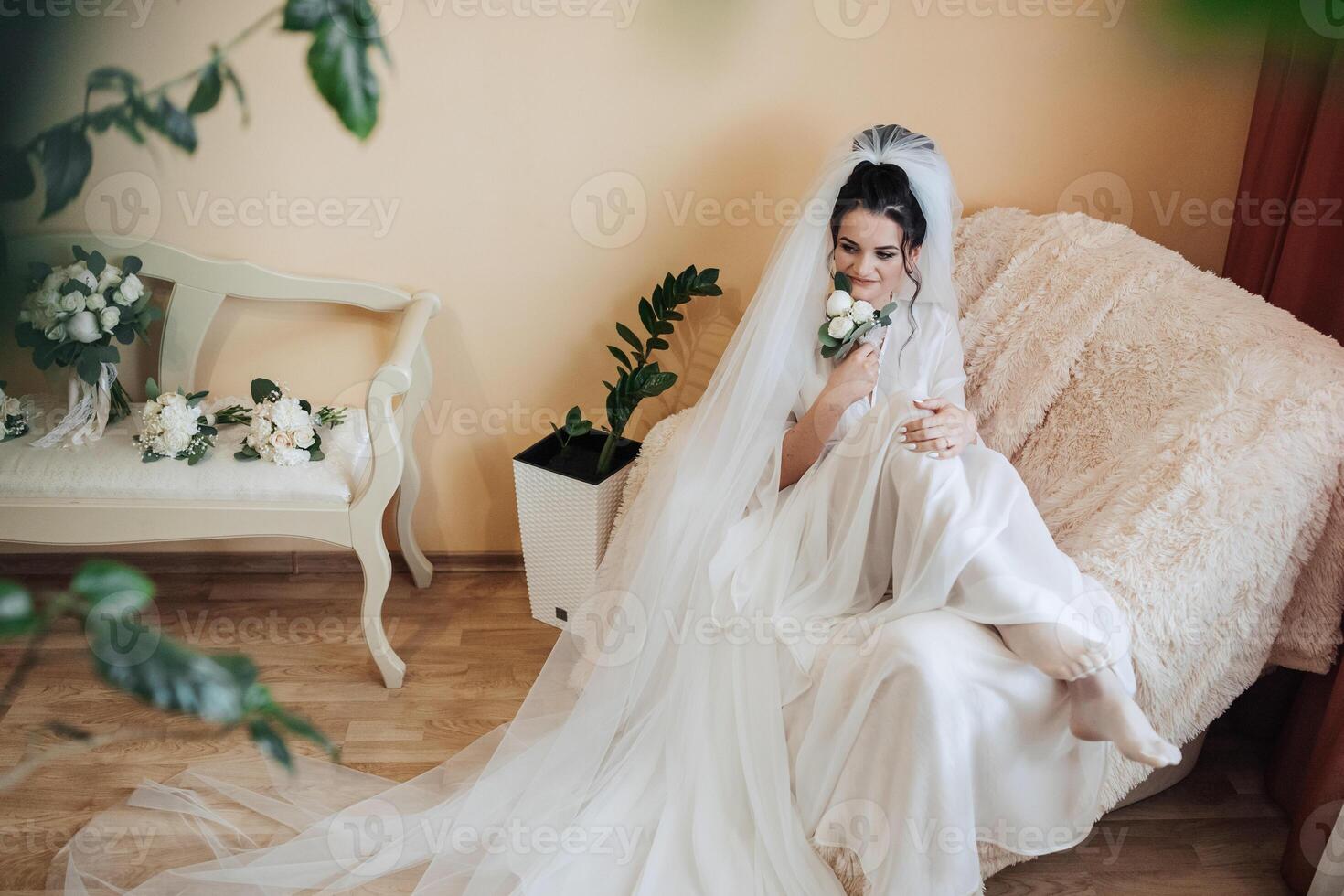 A bride is sitting on a couch with a bouquet of flowers in her lap. She is wearing a white dress and a veil photo