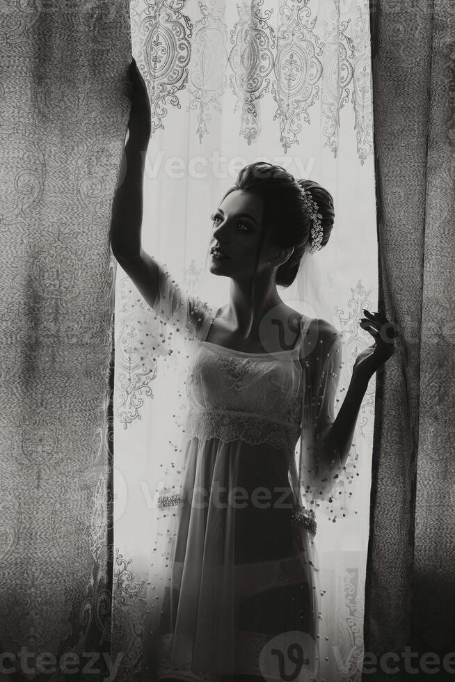 A woman in a wedding dress stands in front of a window. The image is black and white, giving it a classic and timeless feel. The woman's pose and the window's light create a sense of elegance photo