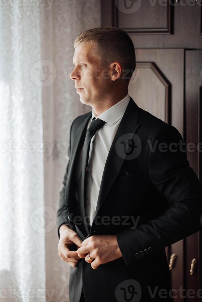 A man in a suit is standing in front of a window, adjusting his tie. Concept of formality and professionalism, as the man is dressed in a suit and tie. The window provides a view of the outside world photo