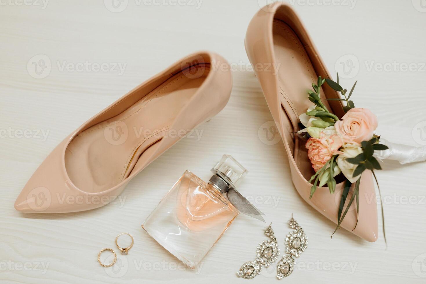 A pair of shoes with a flower on the toe and a bottle of perfume next to them. The perfume bottle is a small square bottle with a pink label photo