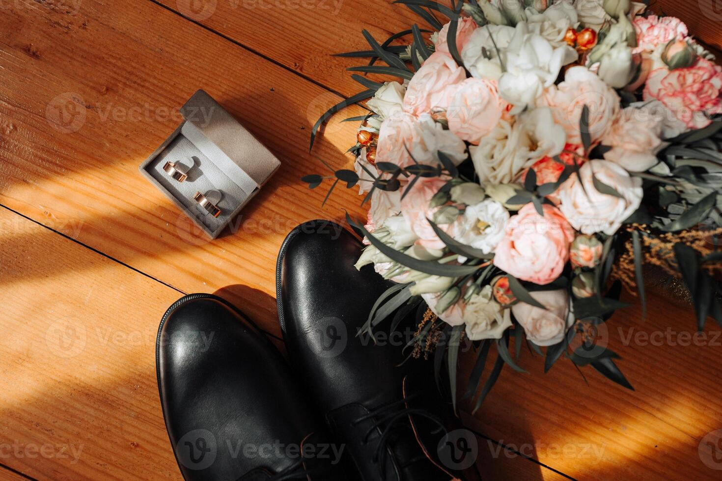 A pair of black shoes and a bouquet of flowers are displayed on a wooden floor. The shoes are placed next to a small box containing a ring and another ring. The flowers are arranged in a vase photo