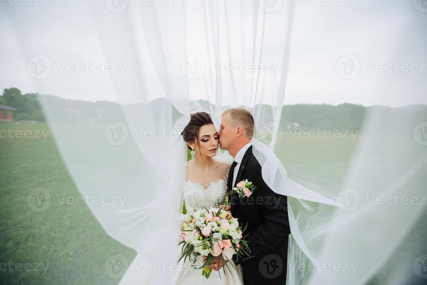 A bride and groom are kissing in front of a white curtain. The bride is wearing a white dress and the groom is wearing a black suit. The scene is intimate and romantic photo