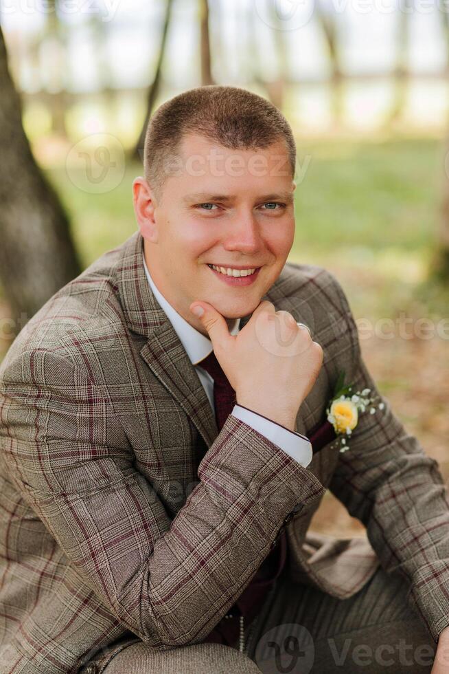 A man in a suit is sitting on the ground with his hands on his knees. He is smiling and looking at the camera photo