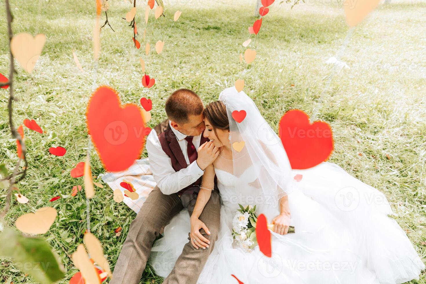 A bride and groom are sitting on a blanket in a field with red hearts surrounding them. The scene is romantic and intimate, with the couple embracing each other photo