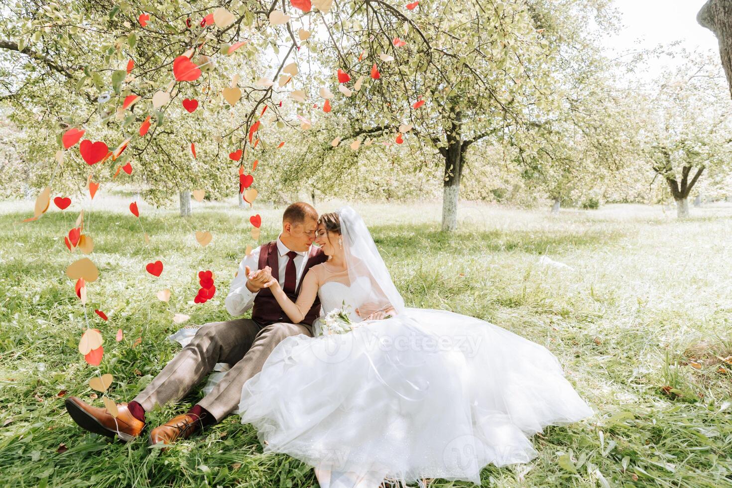 A bride and groom are sitting in a field with red and yellow paper hearts falling around them. The scene is romantic and peaceful photo