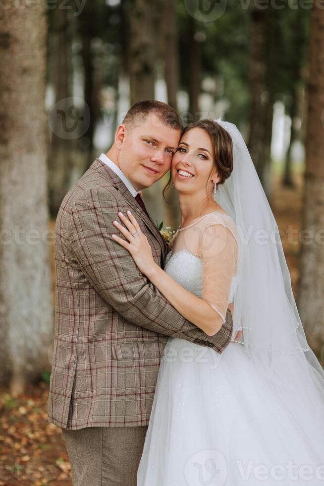 A bride and groom are posing for a picture in a forest. The bride is wearing a white dress and the groom is wearing a suit. They are both smiling and looking at the camera photo